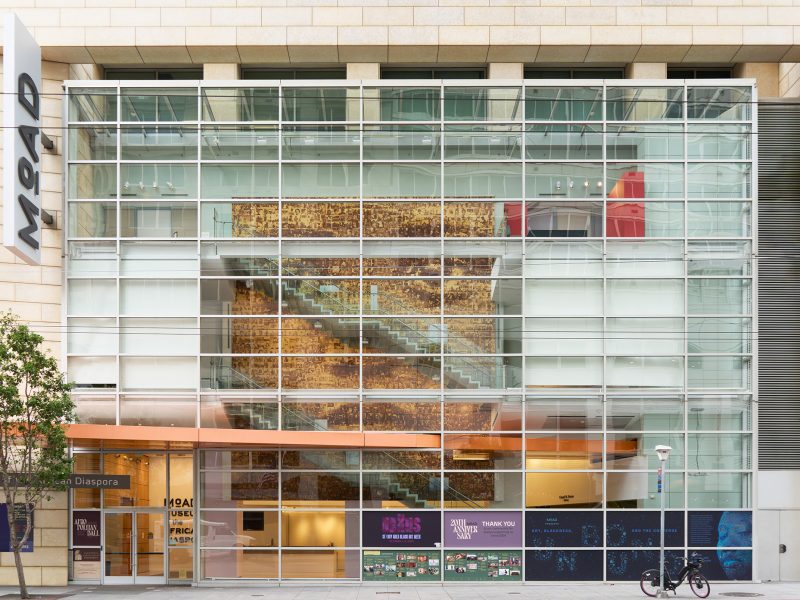 Exterior view of the Museum of the African Diaspora featuring a glass facade, visible staircase, and various colorful posters at the entrance.