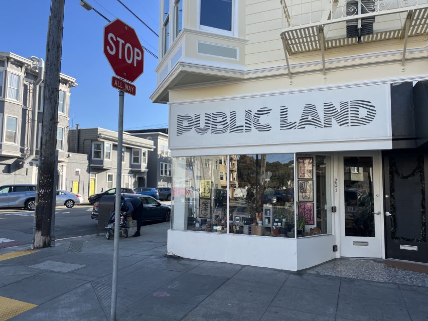 Street corner with a "STOP ALL WAY" sign and a storefront labeled "PUBLIC LAND" displaying framed art in the window.