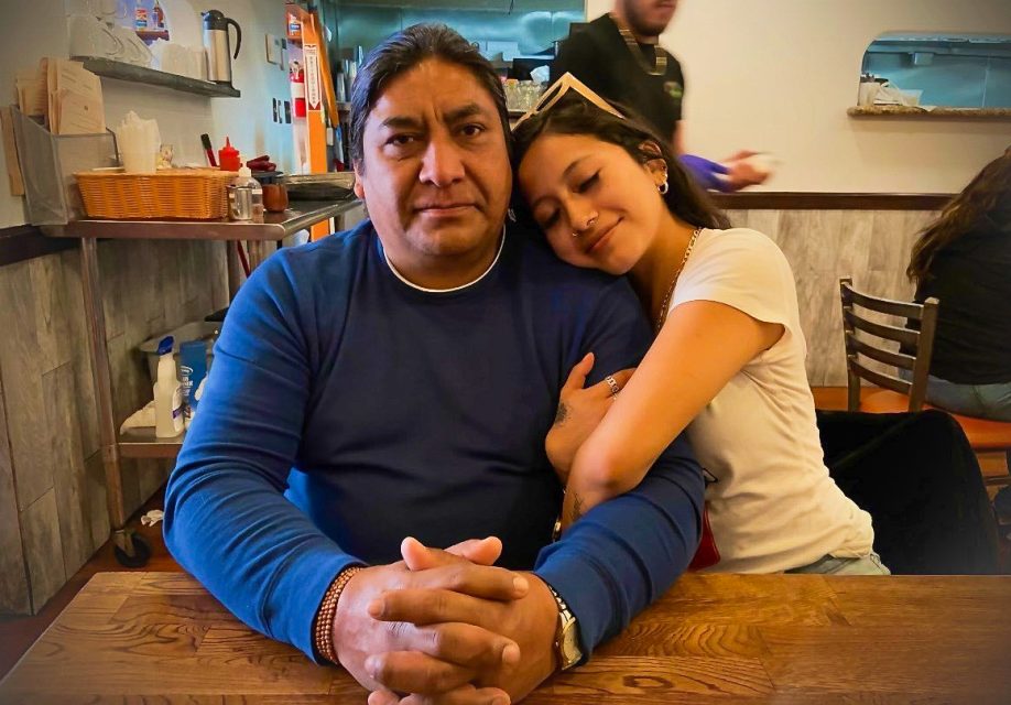 A man sits at a wooden table with his hands clasped while a woman rests her head on his shoulder, both facing the camera inside a casual restaurant.
