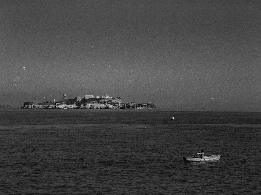 A person rows a small boat in the foreground with Alcatraz Island and its buildings visible in the distance under a clear sky.