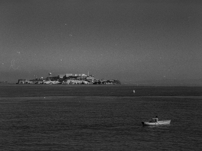 A lone person rows a small boat on calm water with Alcatraz Island and its prison buildings visible in the distant background under a clear sky.