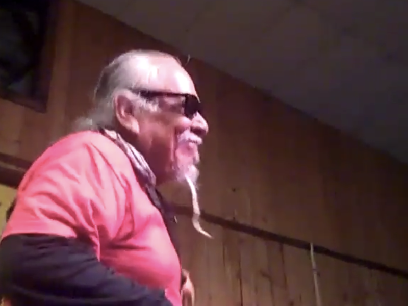 An older man with long white hair, sunglasses, and a goatee wears a red shirt over a black long-sleeve shirt, standing in a wood-paneled room.