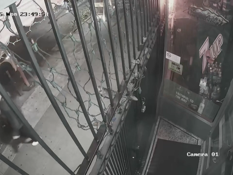 Surveillance camera view of a gated stairwell entrance at night, with a person walking by on the sidewalk, lights above, and a KitKat wrapper visible near the gate. Timestamp reads 10-27-2023, 23:49:31.