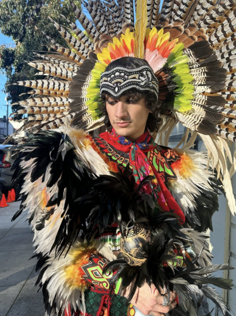 A person wearing a colorful feathered headdress and garment stands outdoors in sunlight, with detailed beadwork and patterns visible on their outfit.