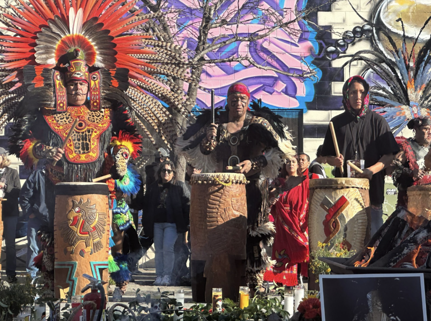 Three men in feathered headdresses and traditional attire play large decorated drums outdoors, with a colorful mural and onlookers in the background.