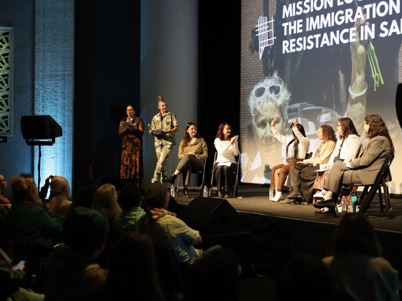 A panel of seven people sits on stage in front of an audience, with a large screen behind them displaying text about immigration policy and crisis resistance in San Francisco.