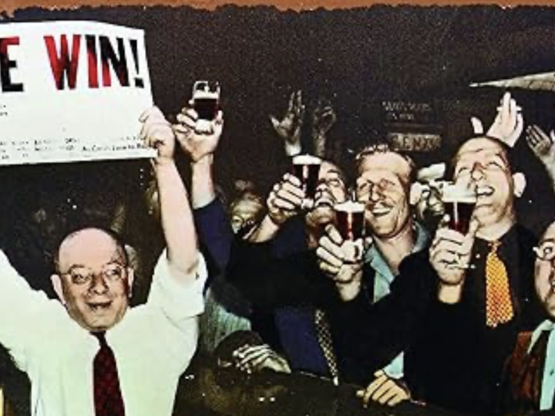 A group of men in suits cheer and raise drinks while one man holds a sign that reads “WE WIN!” in bold letters.