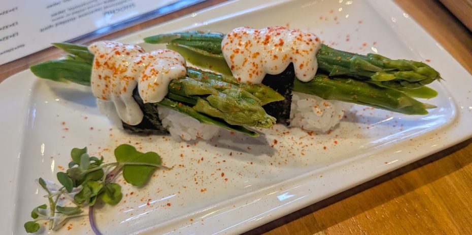 Two pieces of sushi topped with asparagus, creamy sauce, and red seasoning sit on a rectangular white plate, garnished with microgreens.