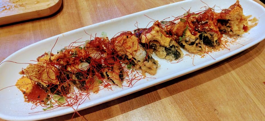 A long white plate with a row of sushi rolls topped with crispy tempura flakes and thin red chili threads, on a wooden table.