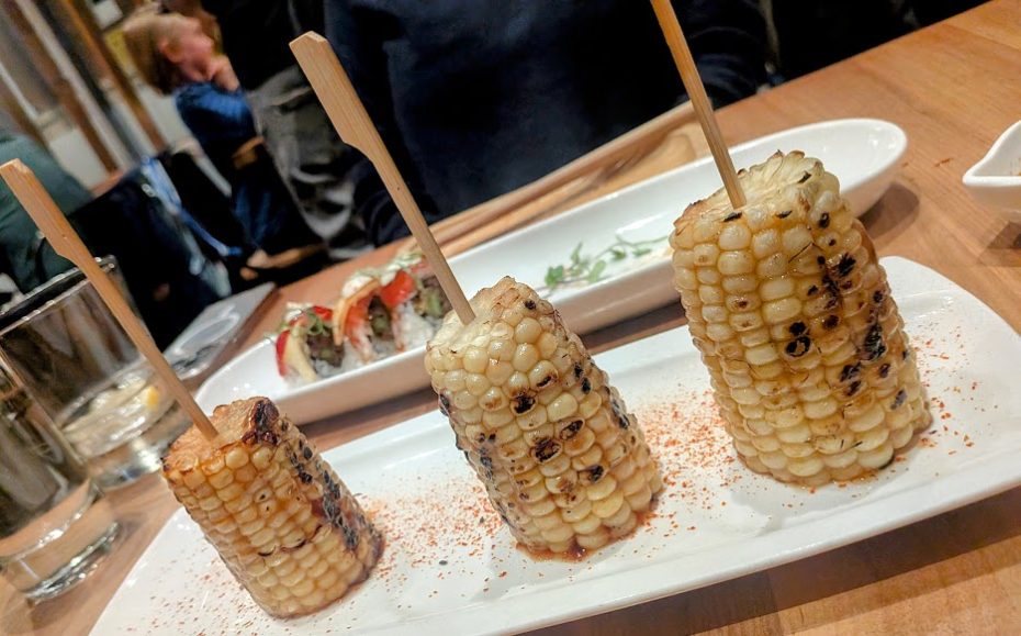 Three grilled corn cobs on sticks are served on a white plate, sprinkled with seasoning. A plate of sushi is visible in the background.