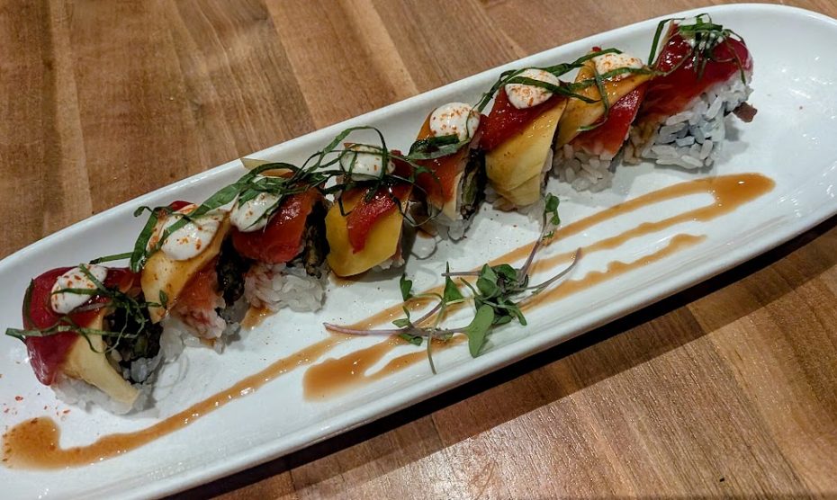 A plate of sushi rolls topped with assorted fish, garnished with herbs and sauce, is arranged on a white rectangular dish on a wooden table.