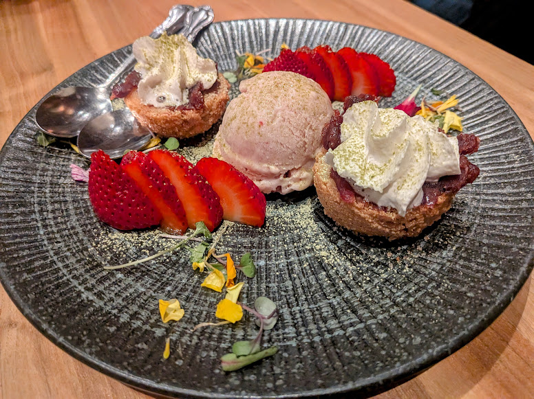 A black plate with two desserts topped with cream, a scoop of ice cream, sliced strawberries, edible flowers, and two spoons.