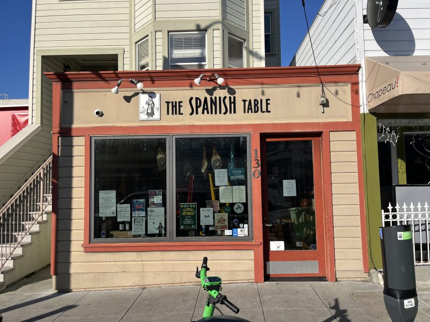 Storefront of The Spanish Table with posters and notices on the window, a red door, tan and red trim, and a green scooter parked on the sidewalk in front.