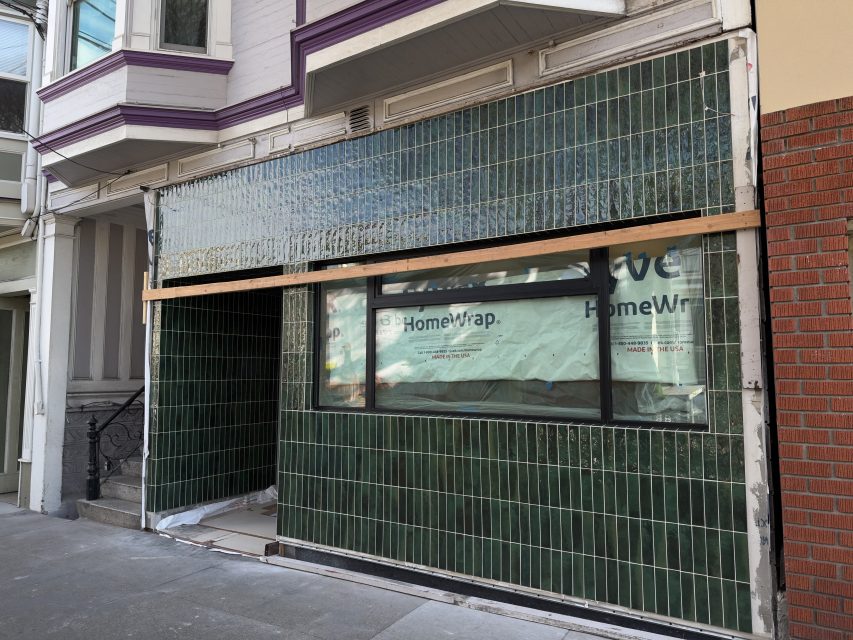 A storefront under renovation features vertical green tiles, a large window covered with HomeWrap, and wooden planks supporting the facade.
