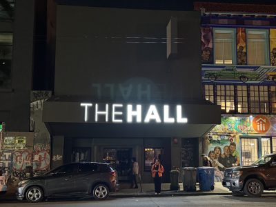 A building at night with "THE HALL" illuminated on its facade; a person in an orange vest stands near parked cars and colorful murals are visible on the adjacent building.