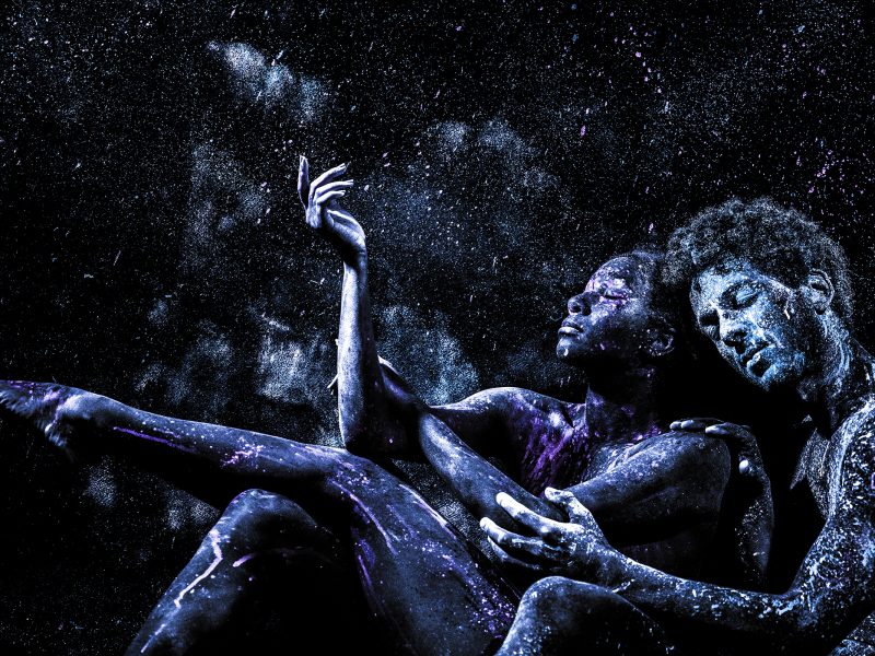Two people with bodies painted in dark blue and purple tones pose closely together against a black background with scattered powder creating a starry effect.