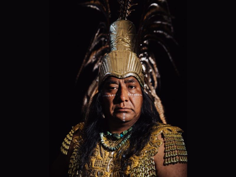 A man wearing elaborate traditional indigenous attire, including a feathered headdress, golden armor, face paint, and beaded necklaces, poses against a dark background.