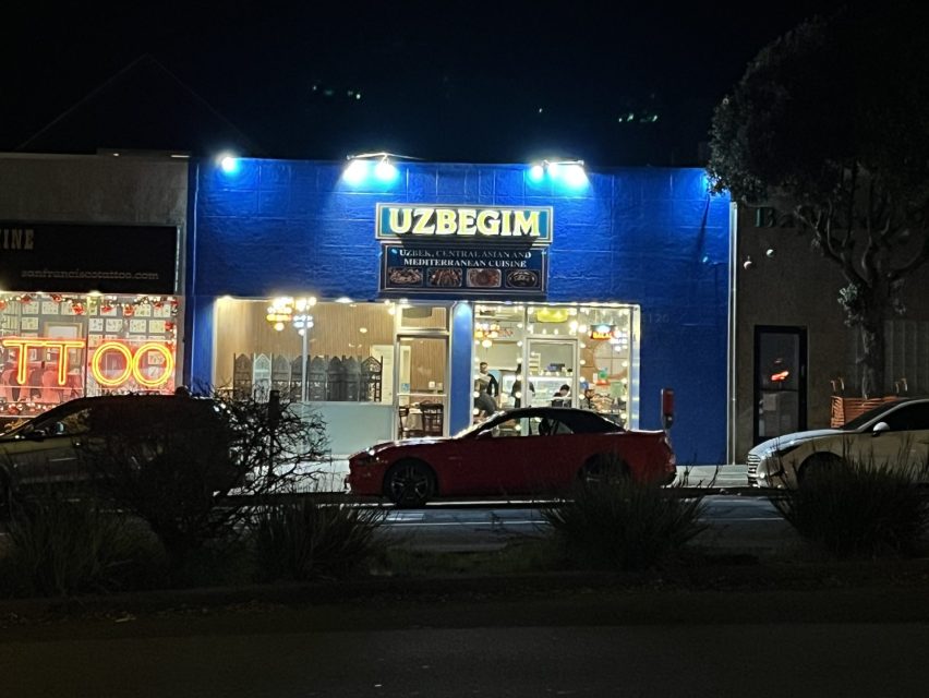 A brightly lit Uzbek restaurant named "UZBEGIM" is open at night, with cars parked in front and several people visible inside.