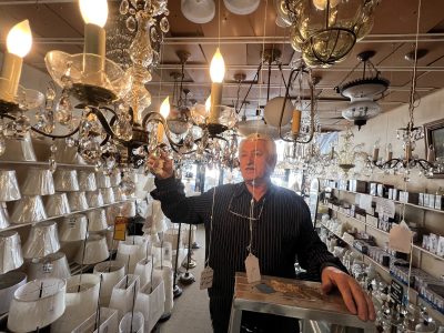 An older man standing on a stepladder adjusts a chandelier in a lighting store filled with lampshades and hanging light fixtures.