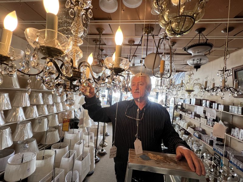 An older man standing on a stepladder adjusts a chandelier in a lighting store filled with lampshades and hanging light fixtures.