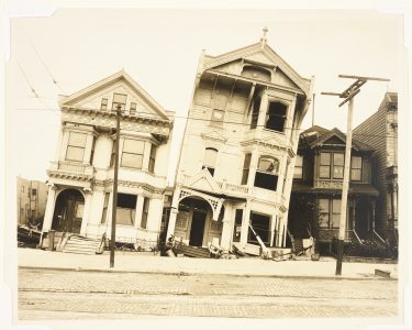 Two damaged Victorian-style houses lean to the side, with broken windows and collapsed porches, on a deserted street lined with cobblestones and utility poles.