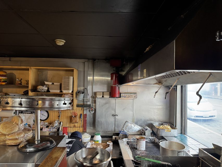 A view of a restaurant kitchen with stainless steel surfaces, shelves with ingredients, a stack of buns, and a window looking out onto a street with a parked car glowing in the warm light of sunset.