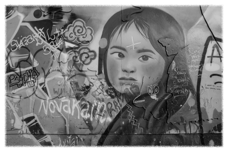 Black-and-white photo of a mural showing a young girl's face, partially covered by graffiti tags and spray-painted text.