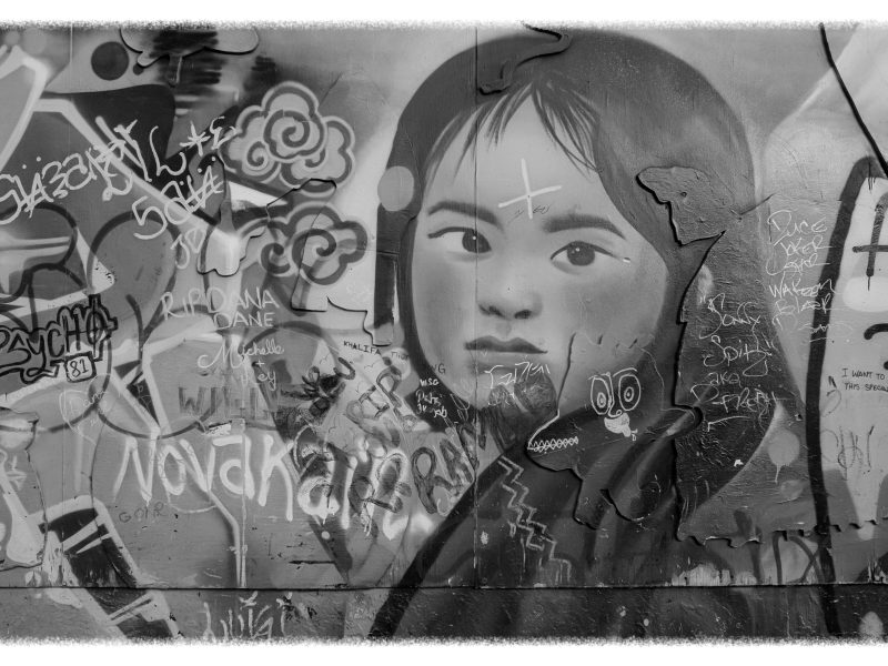 Black-and-white photo of a mural showing a young girl's face, partially covered by graffiti tags and spray-painted text.