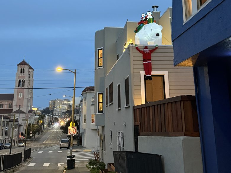 A large inflatable polar bear with two figures on its back and a Santa figure hanging from it is displayed on the second-floor balcony of a house overlooking a quiet city street at dusk.