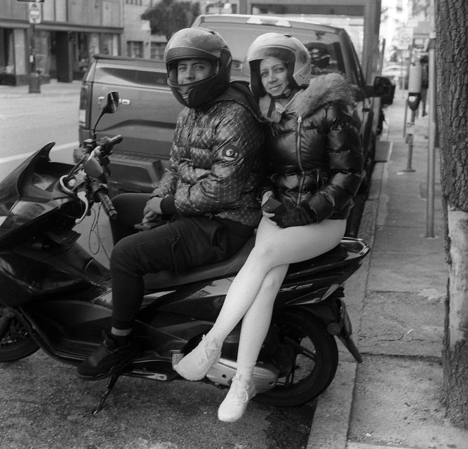 Two people wearing helmets and puffer jackets sit together on a parked scooter by the sidewalk in an urban area.
