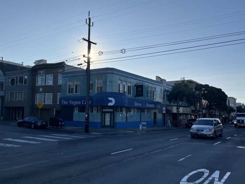 A street corner with a building labeled "The Vegan Cafe" and "Donuts & Donuts" at sunset. Cars are driving by and traffic markings are visible on the road.