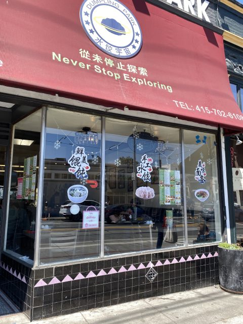 Street view of Dumpling Park restaurant window with food posters, menu, and "Never Stop Exploring" slogan above the entrance. Customers are visible inside.