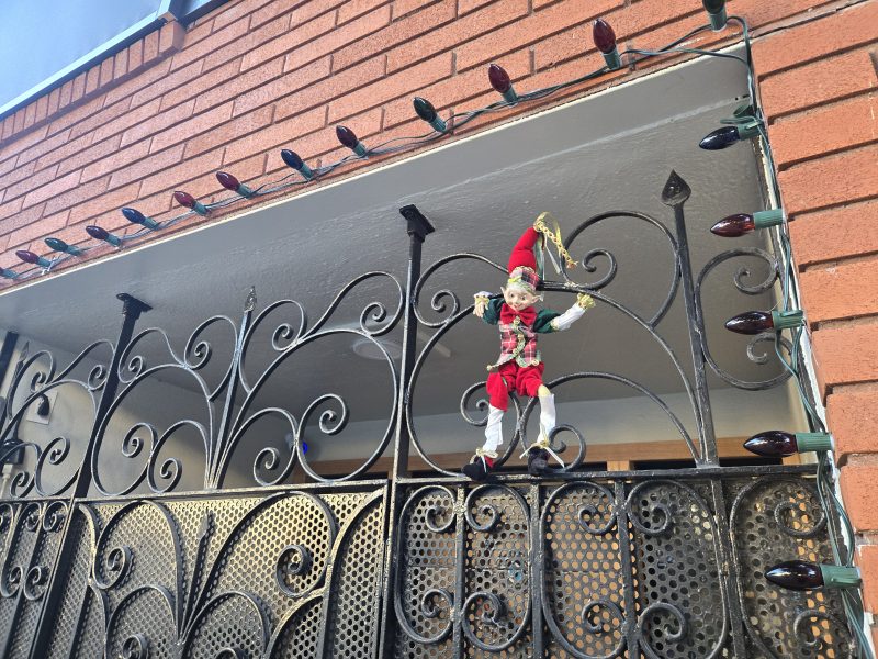 A festive elf doll is hanging on a decorative black metal gate, with Christmas lights strung along the brick wall and balcony above.