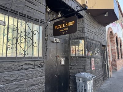 Black storefront with a sign reading "Puzzle Shop est. 2025" above a black door at 3065. Decorative iron bars cover the windows; a trash bin sits on the sidewalk.