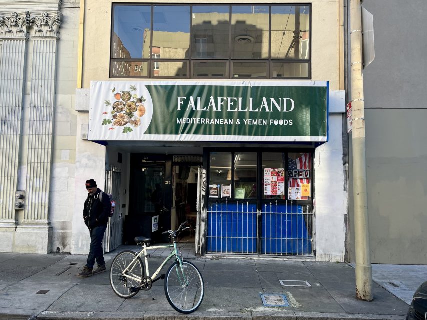 Storefront of Falafelland, a Mediterranean and Yemen foods restaurant, with a bike parked outside and a person standing on the sidewalk.