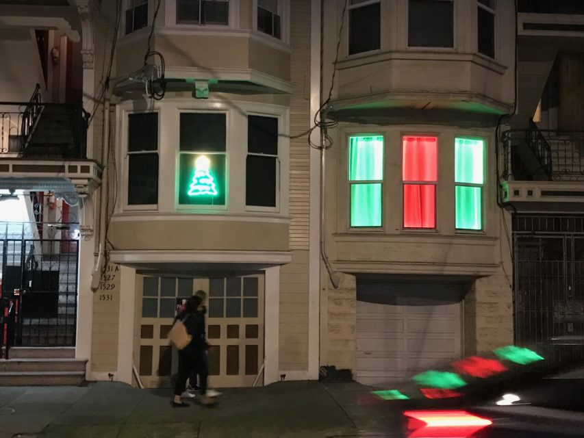 Two people walk past two houses at night, one displaying a neon Christmas tree in the window and the other showing red and green lit curtains.