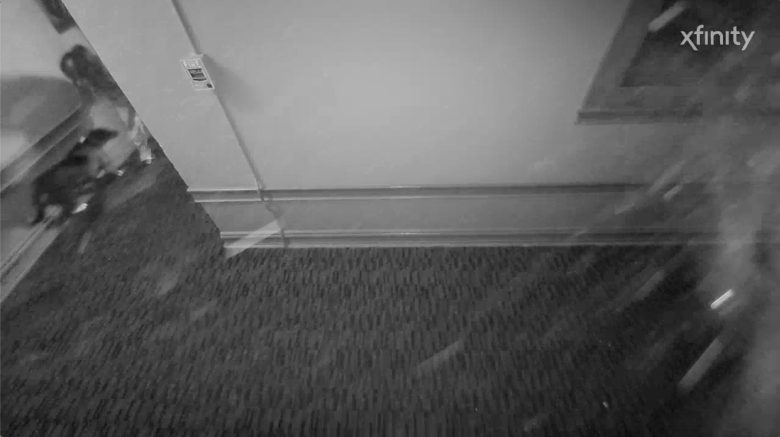 Black and white security camera footage shows a hallway with carpet, a wall outlet, a window, and a person partially visible entering from the left. The Xfinity logo appears in the top right corner.