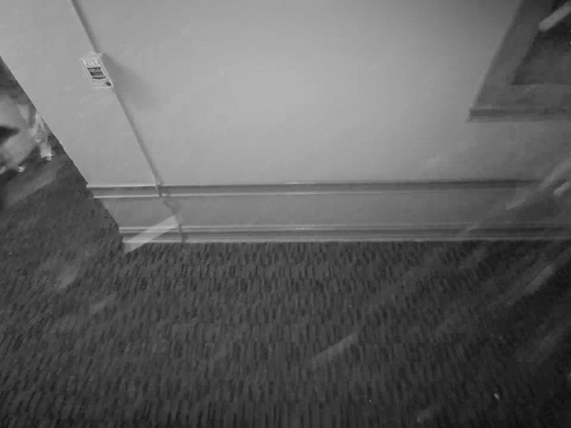 Black and white security camera footage shows a hallway with carpet, a wall outlet, a window, and a person partially visible entering from the left. The Xfinity logo appears in the top right corner.