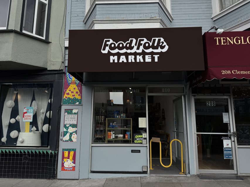 Storefront of Food Folk Market with a brown and white sign, glass door showing "Open" and "Closed" signs, and colorful murals on the left wall.