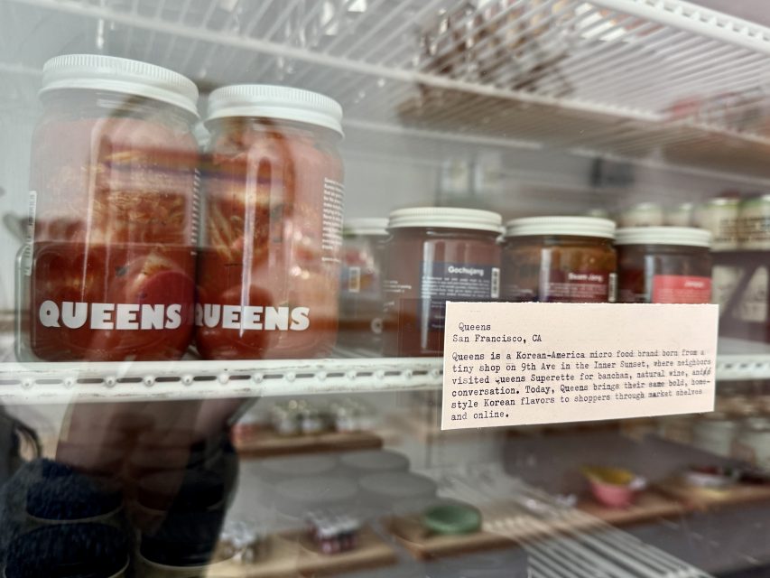 Glass jars labeled "Queens" are displayed on a refrigerator shelf, with a small printed card describing the brand and its products beside them.