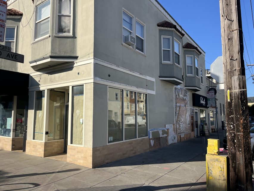A street corner with a beige two-story building featuring empty storefronts, large windows, and various signs; a utility pole and yellow utility boxes are in the foreground.