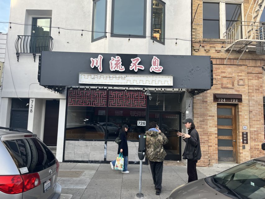 Three people stand outside a closed storefront with Chinese characters on the sign and the street number 726 displayed next to the entrance.