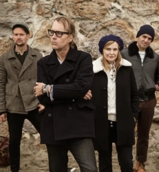 Four adults stand outdoors in front of a stone wall, wearing coats and hats, looking toward the camera with neutral expressions.