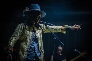 A musician wearing a gold jacket and black hat sings into a microphone and points, performing on a dimly lit stage.