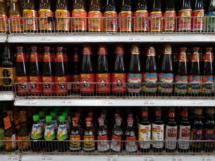 Bottles of various sauces and condiments are displayed on metal shelves in a supermarket aisle.