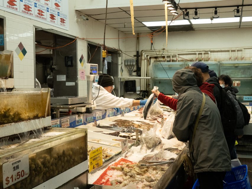 Customers shop for seafood at a market counter, interacting with a vendor behind a display of fish and shellfish on ice.