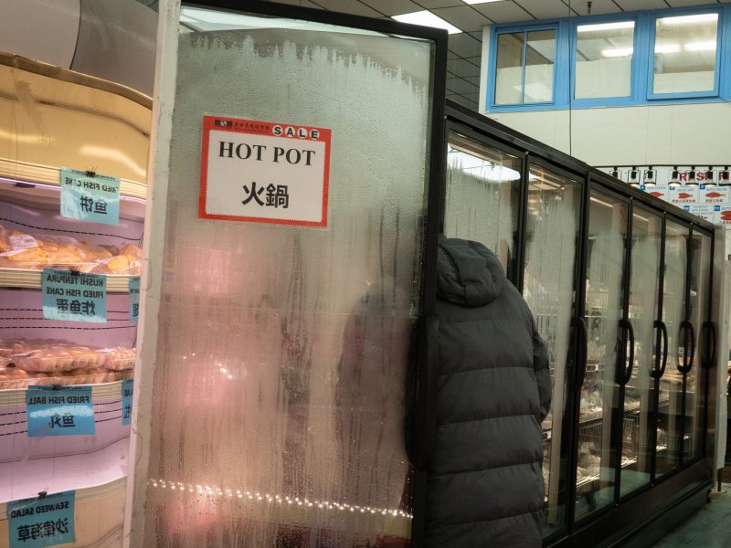 A person wearing a dark coat stands partly inside a foggy glass freezer door labeled "HOT POT" in a grocery store. Shelves of packaged food are visible to the left.