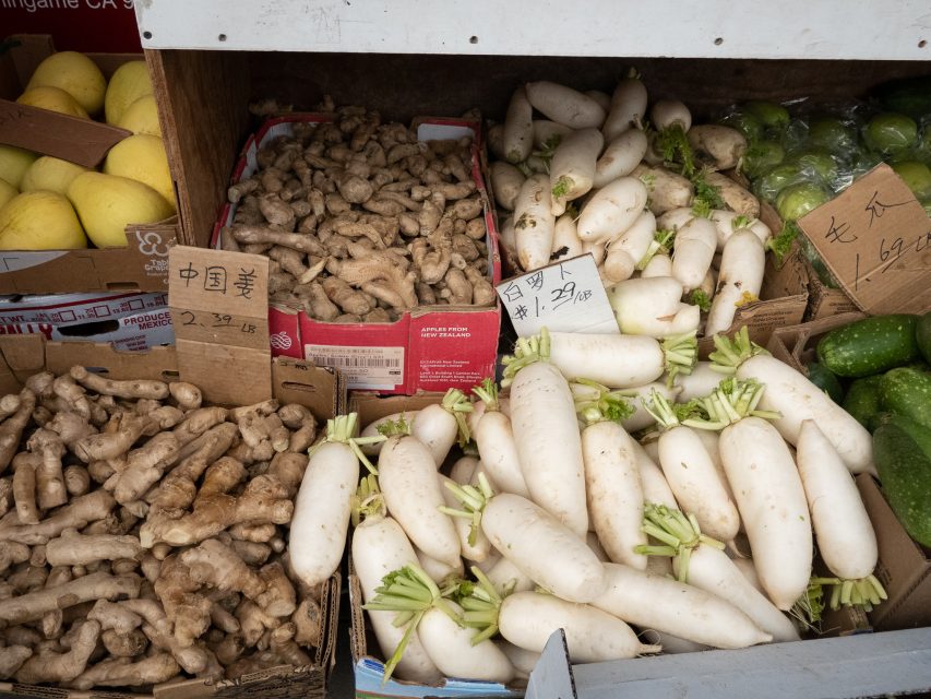 Boxes of fresh ginger roots, white radishes, green bell peppers, and yellow melons displayed for sale at a market, with handwritten price signs in Chinese.
