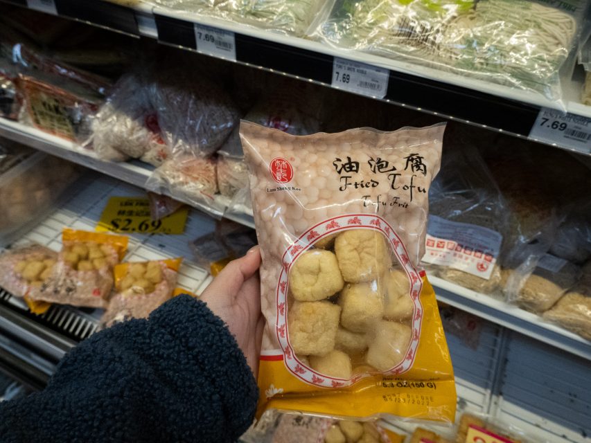 A hand holding a packaged bag of fried tofu in front of a refrigerated grocery store shelf with other tofu products.