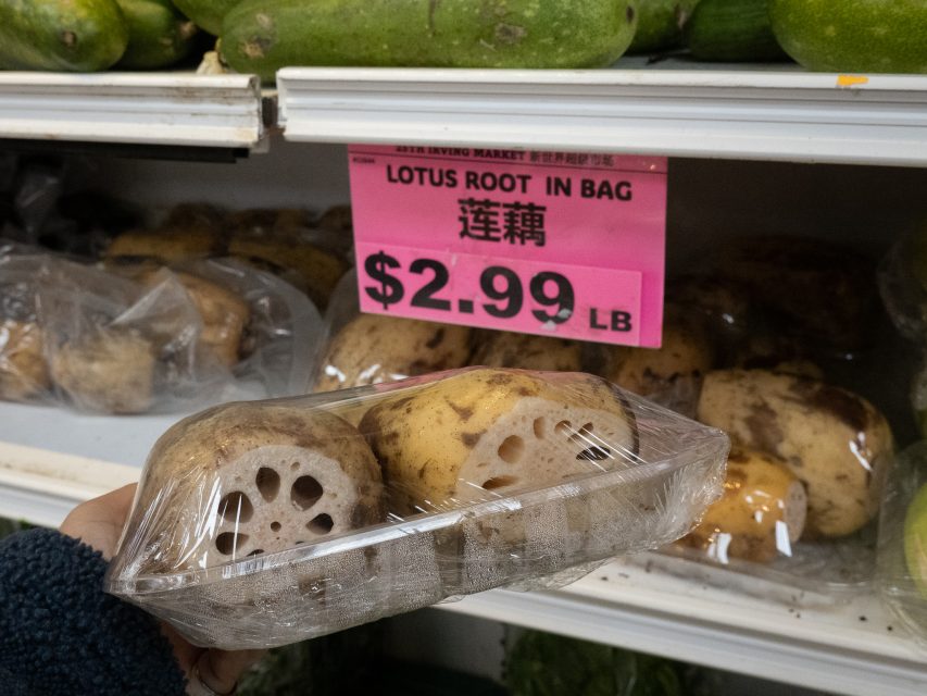 A hand holds a plastic-wrapped package of sliced lotus root in front of a store shelf with a pink sign reading "Lotus Root in Bag $2.99 LB".